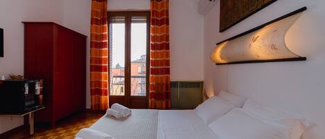 Une chambre d’hôtel avec un grand lit, une armoire rouge, une télévision et une vue sur les immeubles par la fenêtre.