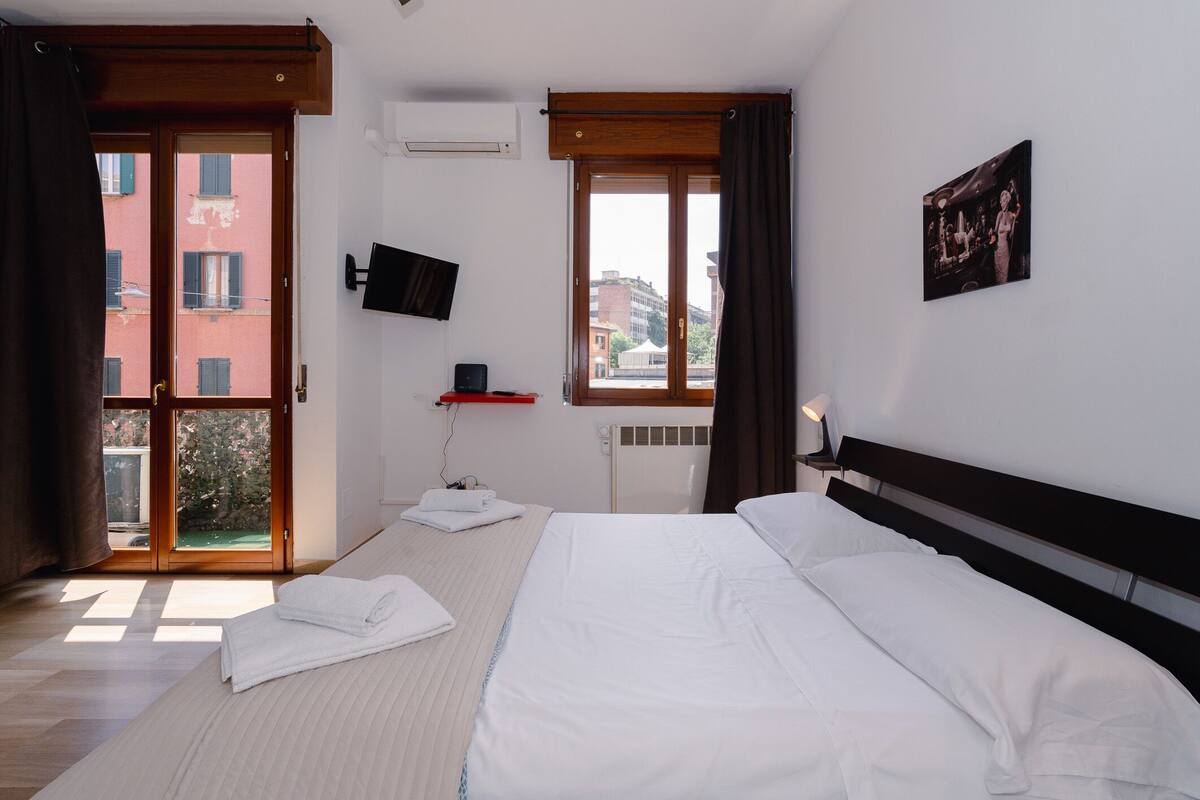 Habitación de hotel con cama, televisor montado en la pared, ventana con cortinas y vistas a edificios.
