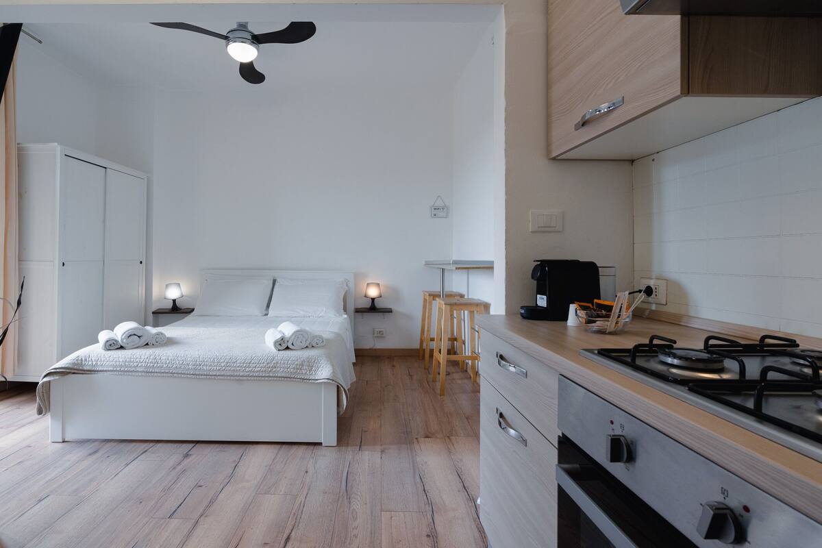 Un dormitorio con cama, lámparas de noche, piso de madera, ventilador de techo y una kitchenette con estufa, además de una pequeña mesa.