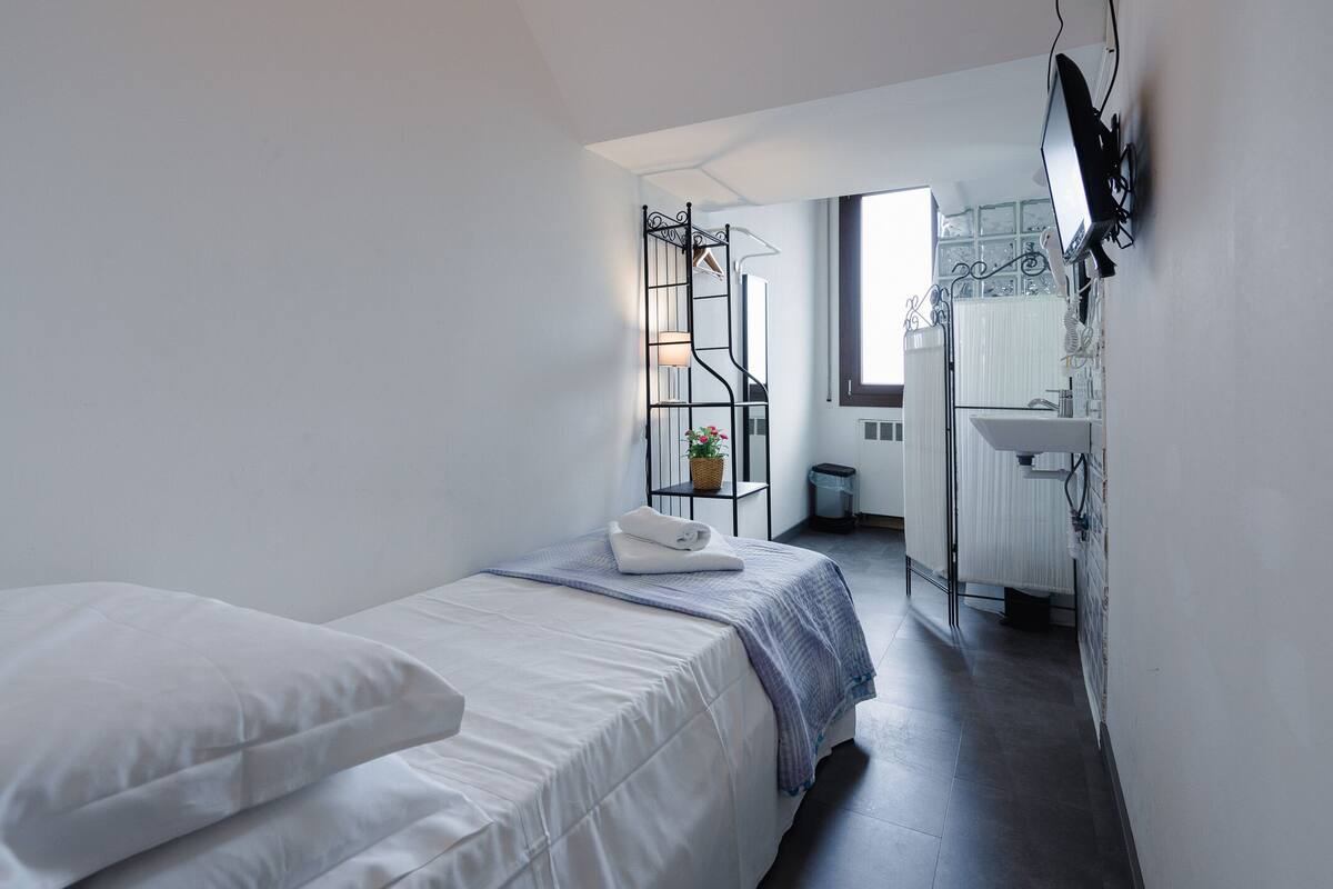 Un dormitorio con cama, una mesita de noche con lámpara, ventana y baño con lavabo y espejo.
