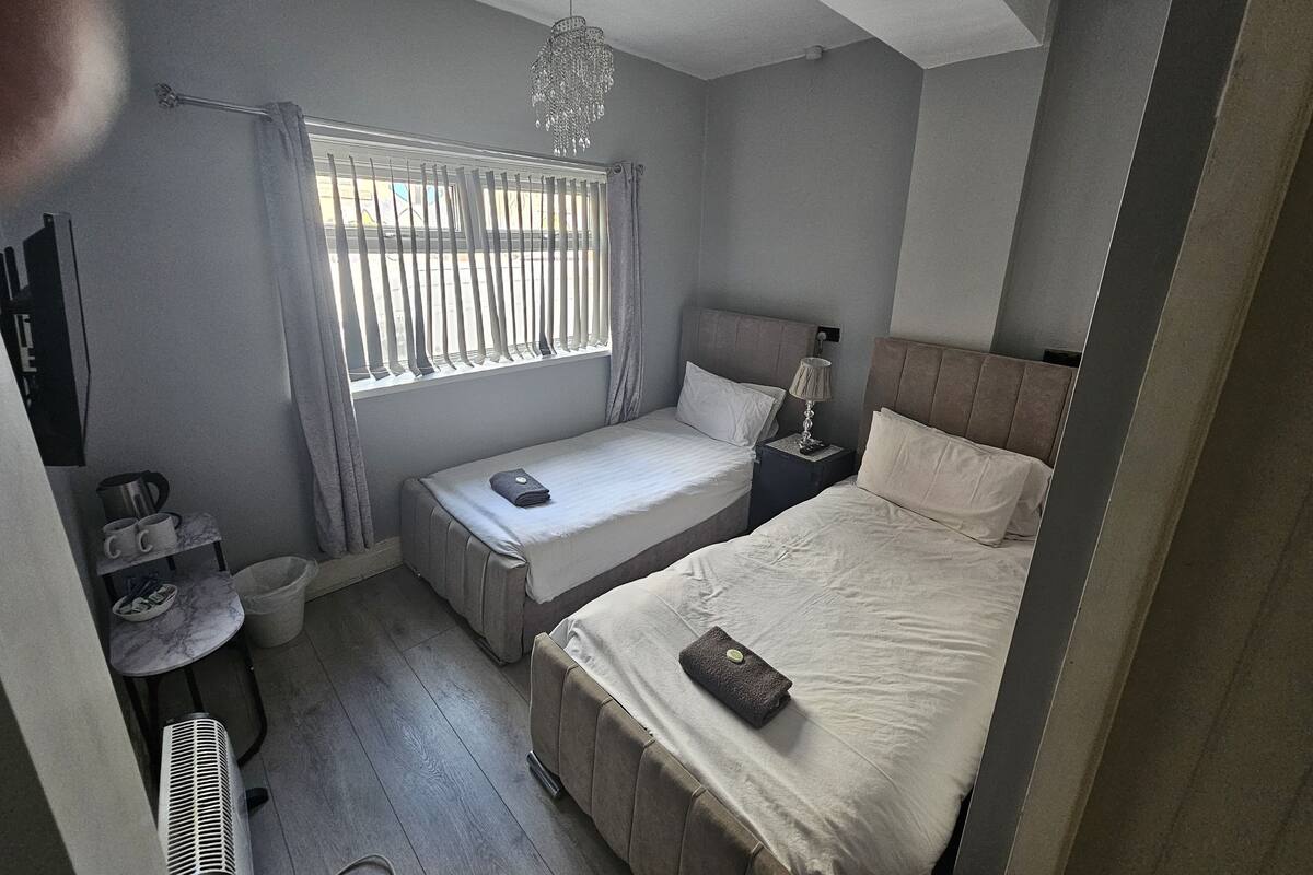 Un dormitorio con una cama individual, una cama más pequeña, una mesita de noche, una lámpara, una ventana con persianas, un calentador y una pequeña mesa con tazas.