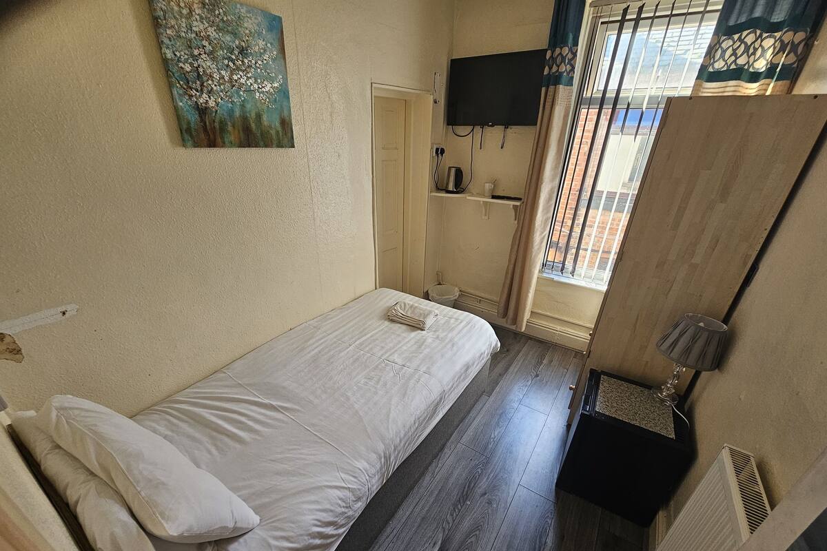 Una habitación pequeña y limpia con una cama individual, un televisor montado en la pared y una ventana que ofrece vistas a edificios.