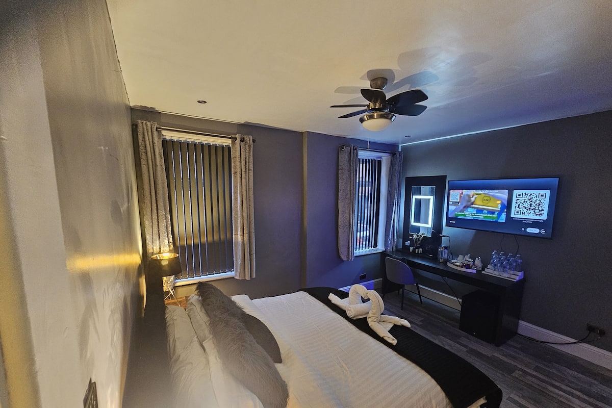 Habitación de hotel con cama, escritorio, computadora, televisor, ventilador de techo y ventana con cortinas.