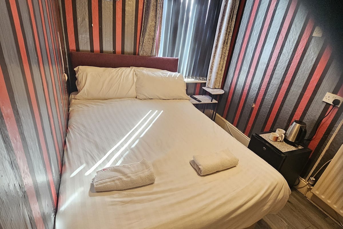 Una cama con colchón blanco y dos almohadas, una mesita de noche con una taza y una tetera, y una ventana con cortinas.
