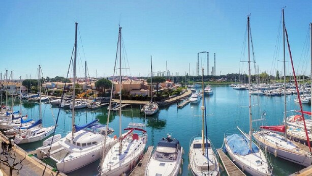 Una marina con diverse imbarcazioni ormeggiate, cielo sereno e edifici sullo sfondo.