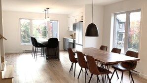 A spacious kitchen with modern appliances, a dining area with a large wooden table and chairs, and large windows offering views of the outside.