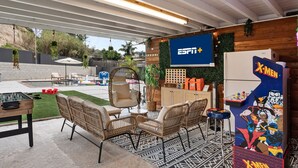 An outdoor patio area with a foosball table, wicker furniture, a pool table, and a large screen displaying ESPN+.
