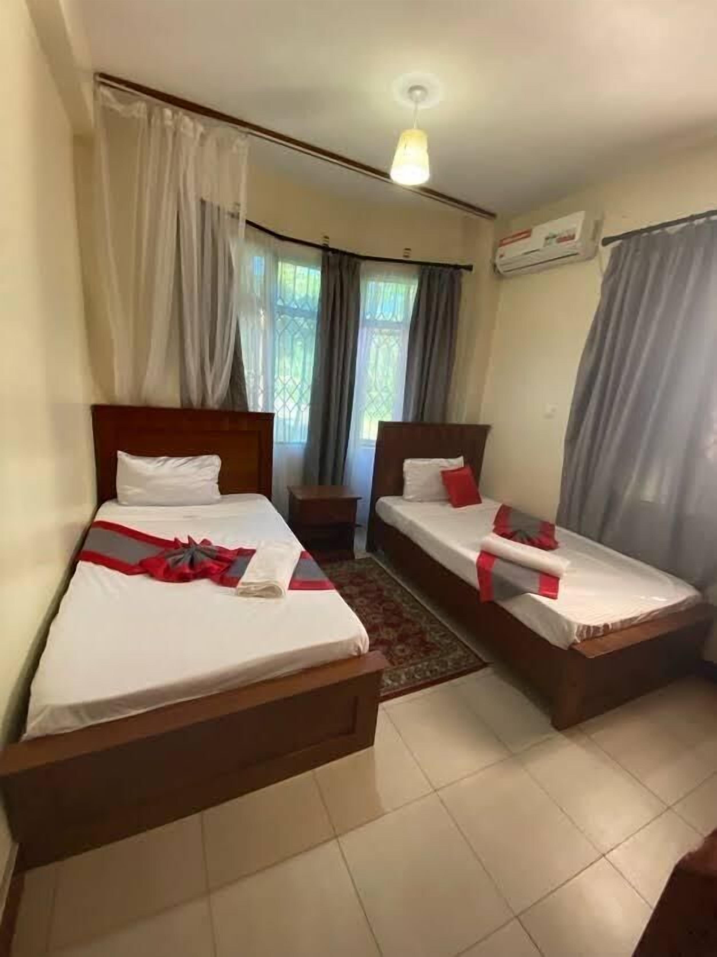 A hotel room with two single beds, a small table, a nightstand, a window with curtains, and an air conditioning unit.
