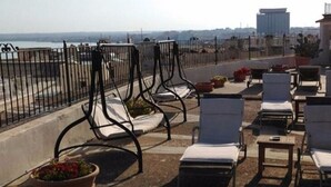 A rooftop terrace with sun loungers and potted plants, overlooking a cityscape.