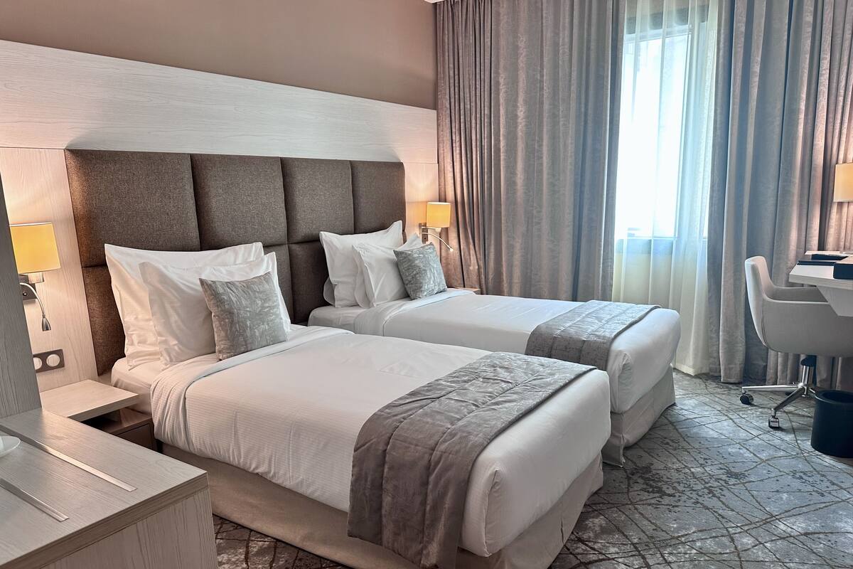 A hotel room with two beds, a desk, a chair, and a large window with curtains.