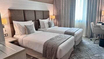 Habitación de hotel con dos camas, un escritorio, una silla y un gran ventanal con cortinas.