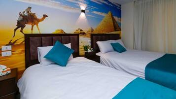 A hotel room with two beds, each with a blue pillow, a mural of a desert scene with pyramids, and a night light.