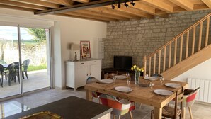 Uma sala de jantar com uma mesa de madeira posta para seis pessoas, uma televisão sobre um suporte e uma parede de pedra ao fundo.