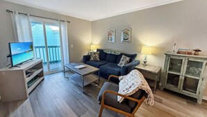 Un salon comprenant un canapé, une table basse, un téléviseur posé sur un meuble, un fauteuil avec une couverture, un petit meuble de rangement et un balcon offrant une vue sur l’océan.