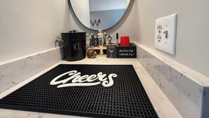 Een badkamer met een zwarte mat op de vloer, een spiegel, een wastafel en een aanrecht met diverse items, waaronder een zwarte emmer met de tekst 'ICE', een doos cocktailglazen en een rode beker.