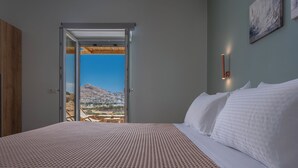 A bedroom with a large bed, a view through an open door, and a wall-mounted light fixture. - Calma Ios (Ios)