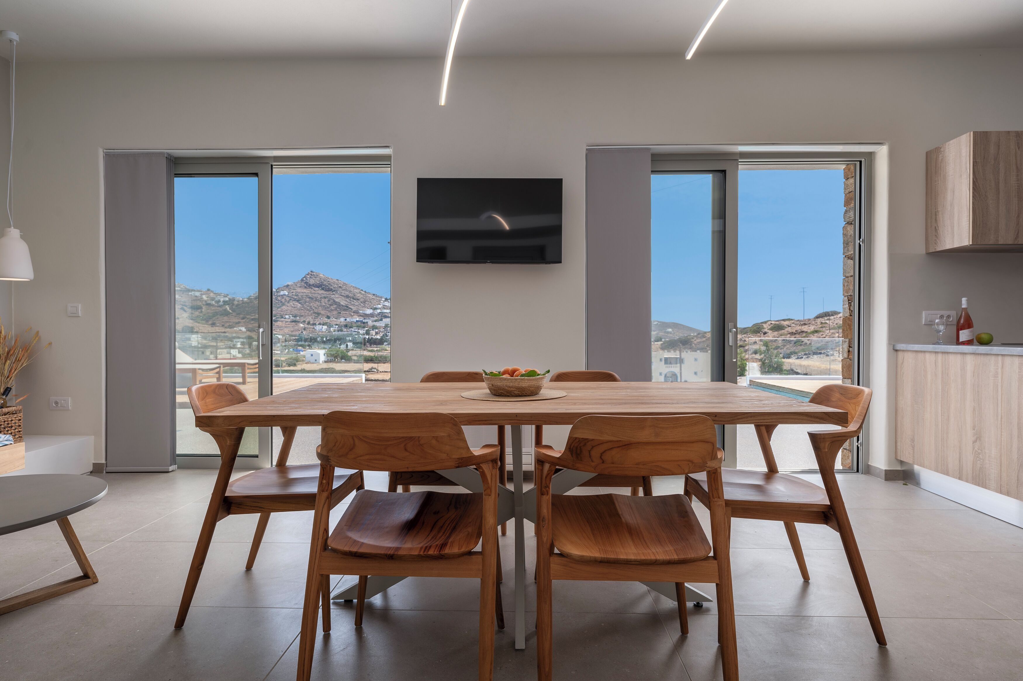 Un'area pranzo moderna con un grande tavolo di legno, sei sedie e una ciotola di frutta. Una TV a schermo piatto è montata sulla parete sopra il tavolo.