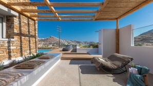 A modern outdoor seating area with a stone wall, a wooden pergola, and a view of the landscape. - Calma Ios (Ios)