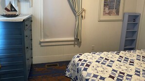 Une chambre à coucher comprenant un lit, une commode, une fenêtre avec des rideaux, un tableau encadré accroché au mur et une petite étagère.