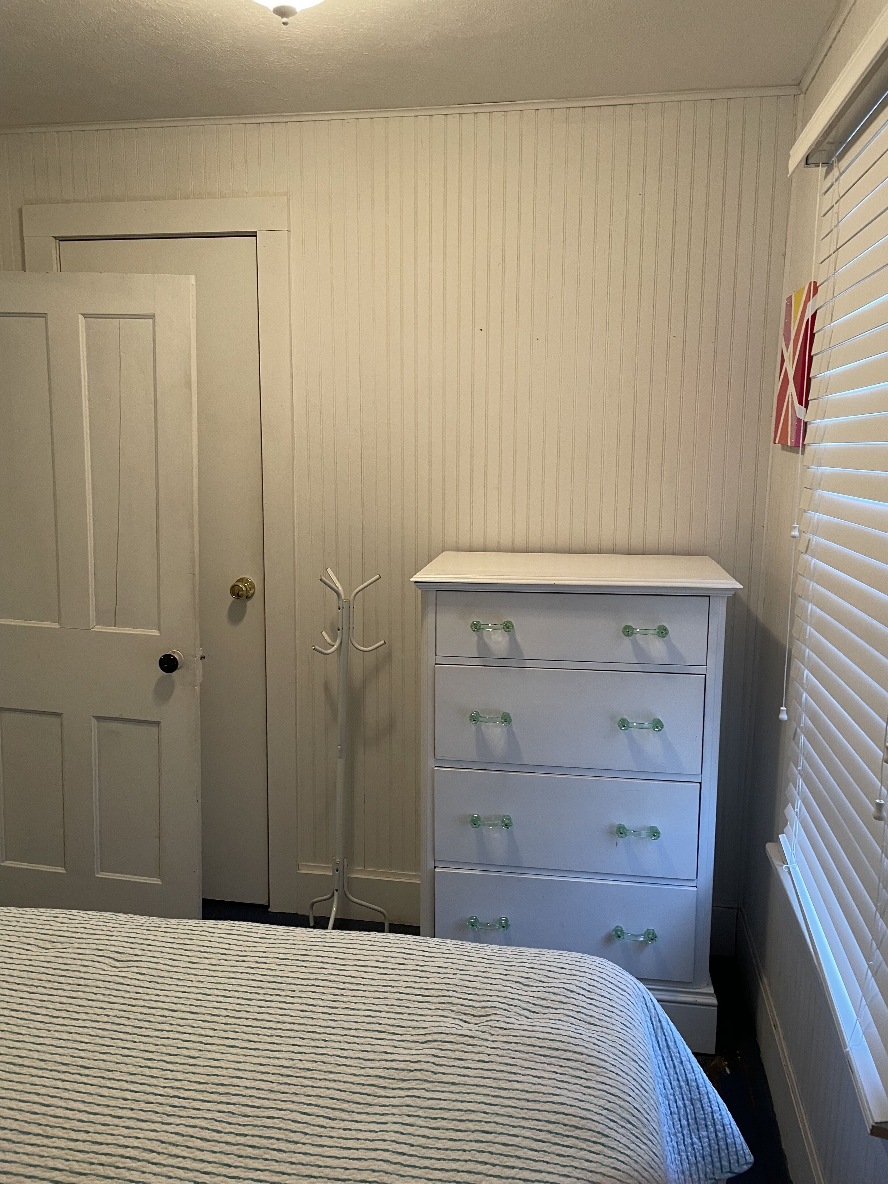A bedroom with a bed, a white dresser with drawers, a coat rack, and a door with a peephole.