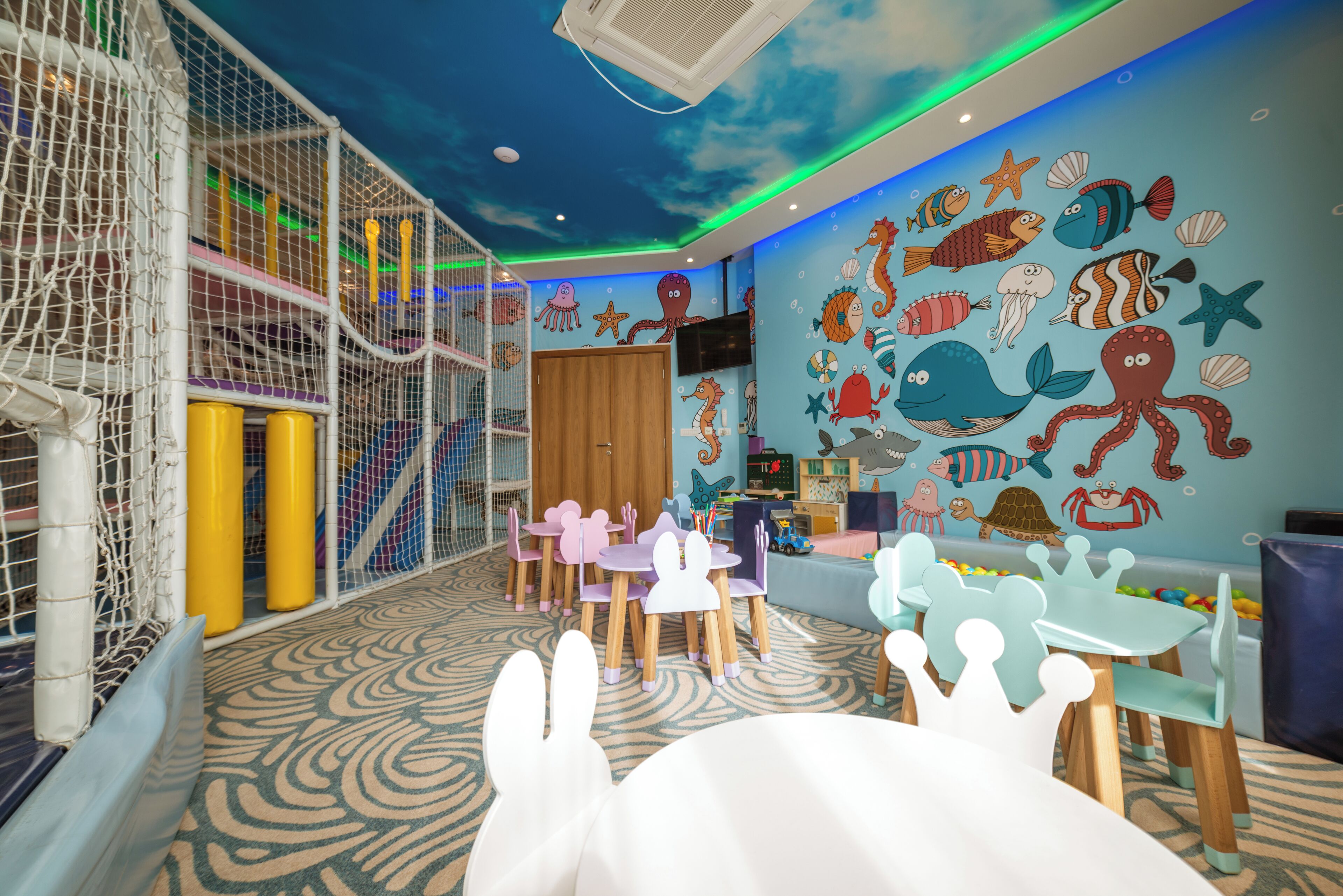 A children's play area with a netted climbing structure, a play table, and a wall decorated with sea creature murals.