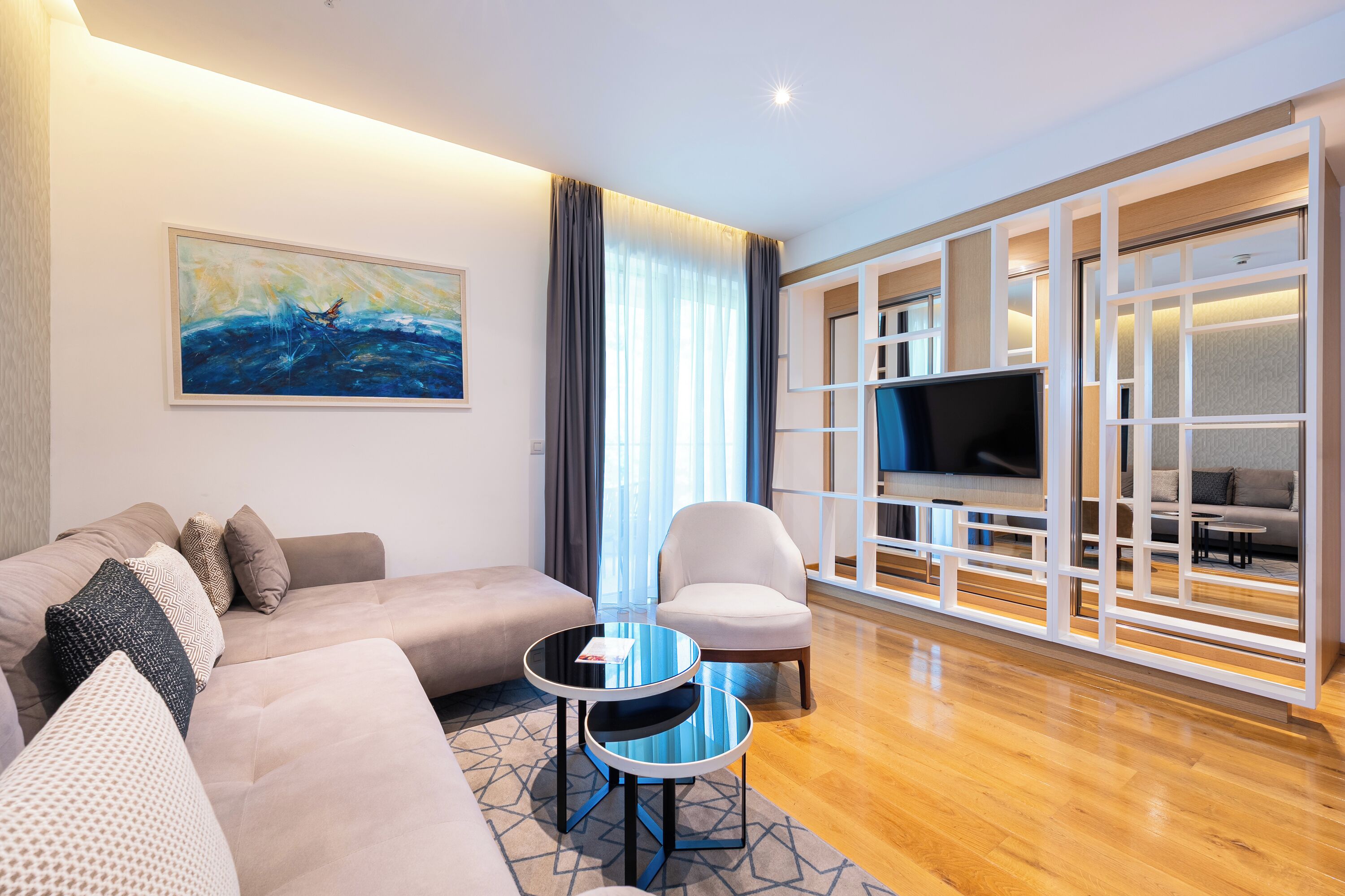 A modern living room with a sofa, armchairs, a coffee table, a TV, and a large window with curtains.