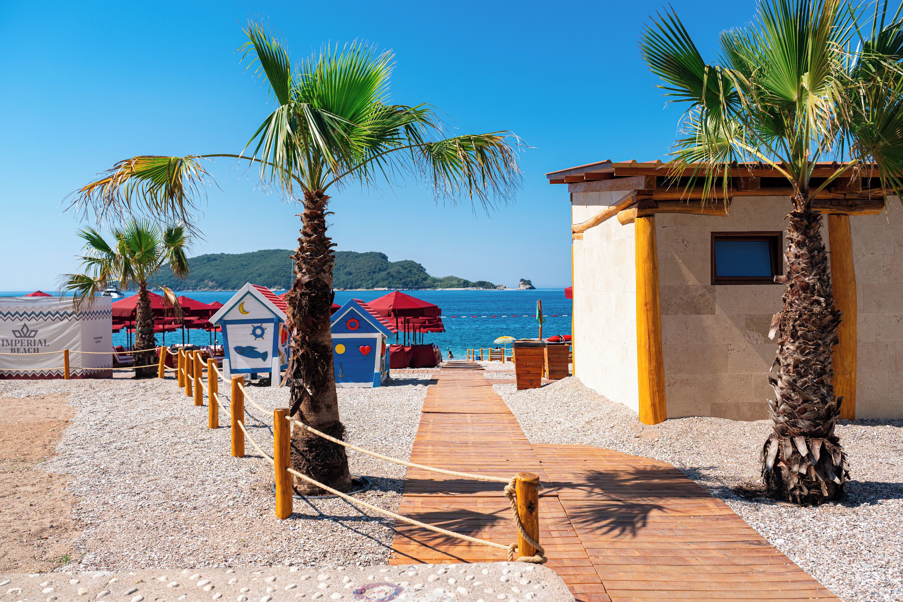 A wooden pathway leading to beach huts with palm trees, a clear blue sky, and the sea in the background.