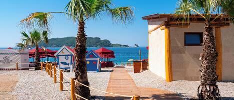 A wooden pathway leading to beach huts with palm trees, a clear blue sky, and the sea in the background.