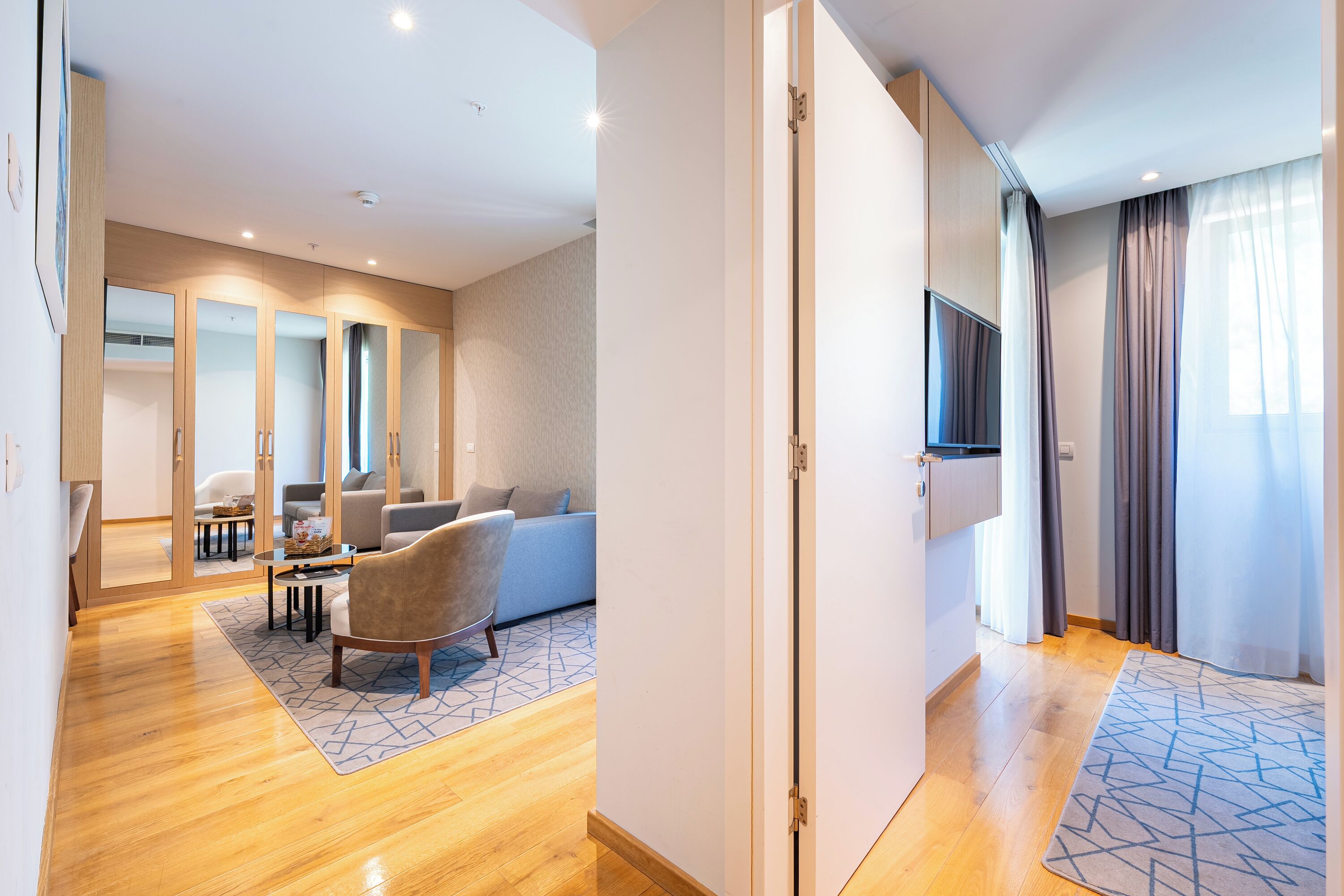 Een moderne hotelkamer met een houten vloer, een bank, een stoel, een televisie en een deur naar een andere kamer.