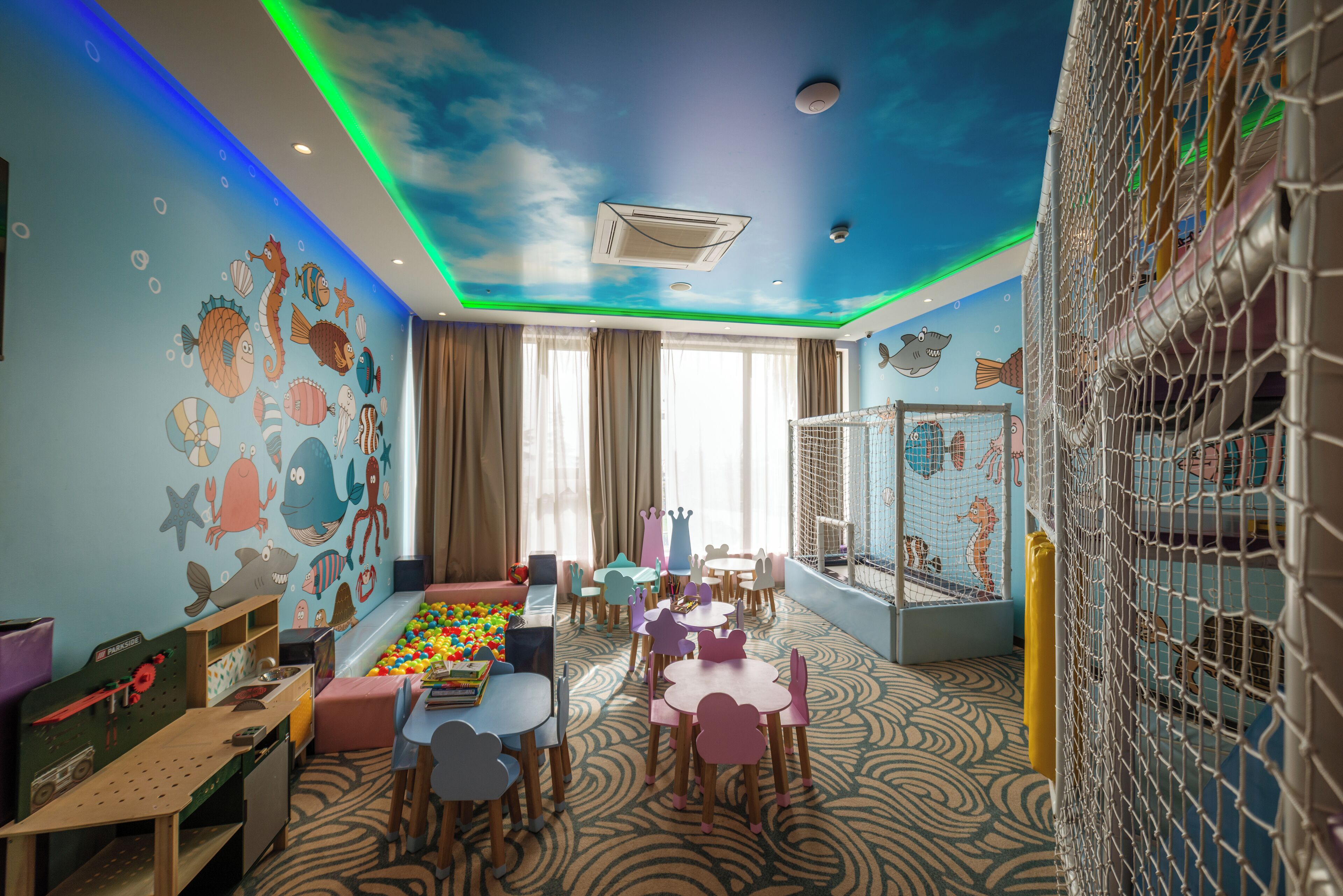 A children's playroom with a ceiling mural, play tables, and a ball pit.