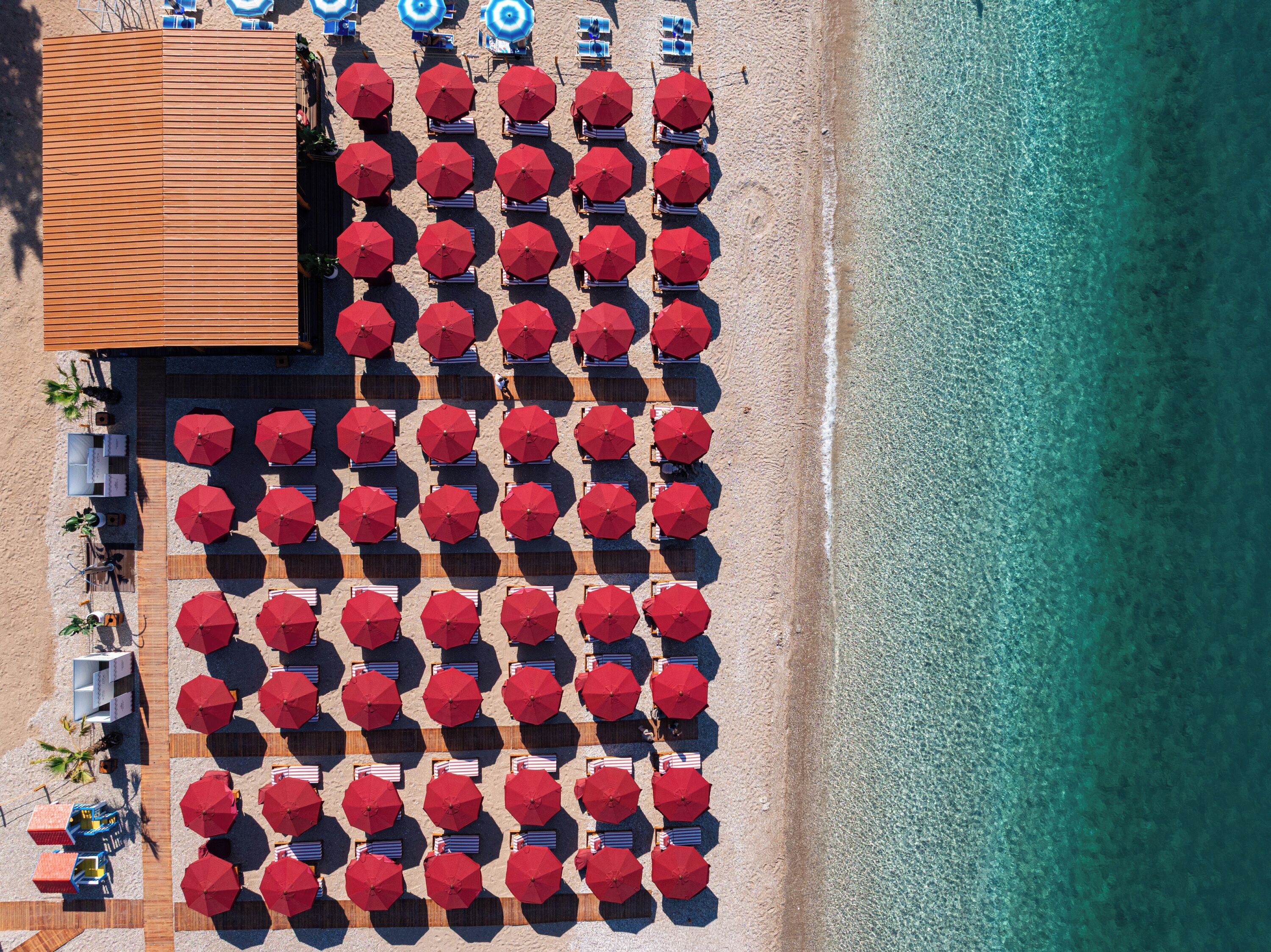 Vista aérea de una playa con hileras de sombrillas rojas y tumbonas, una terraza de madera y un mar turquesa transparente.
