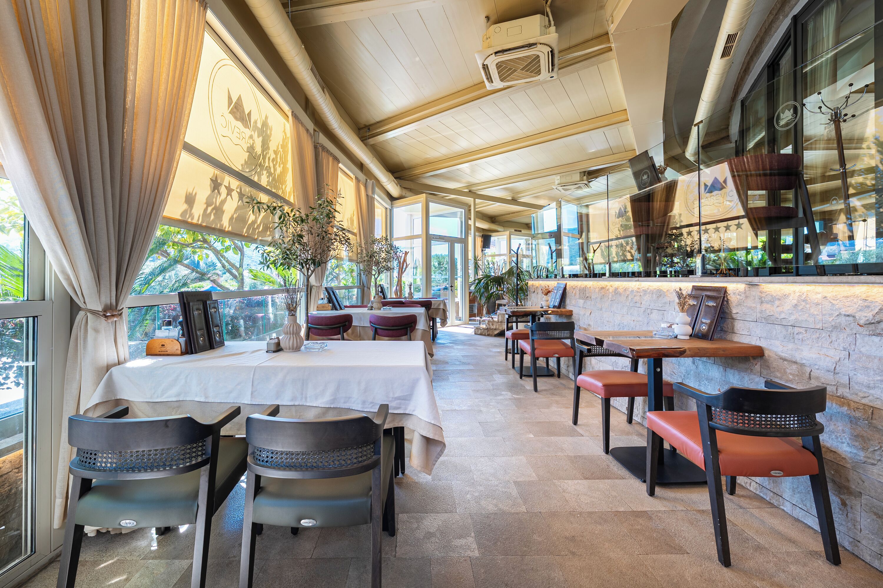A restaurant with tables and chairs, large windows, and a stone wall.