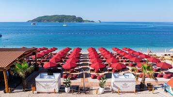 A beachside hotel with rows of red umbrellas, lounge chairs, and a view of the sea and a distant island.