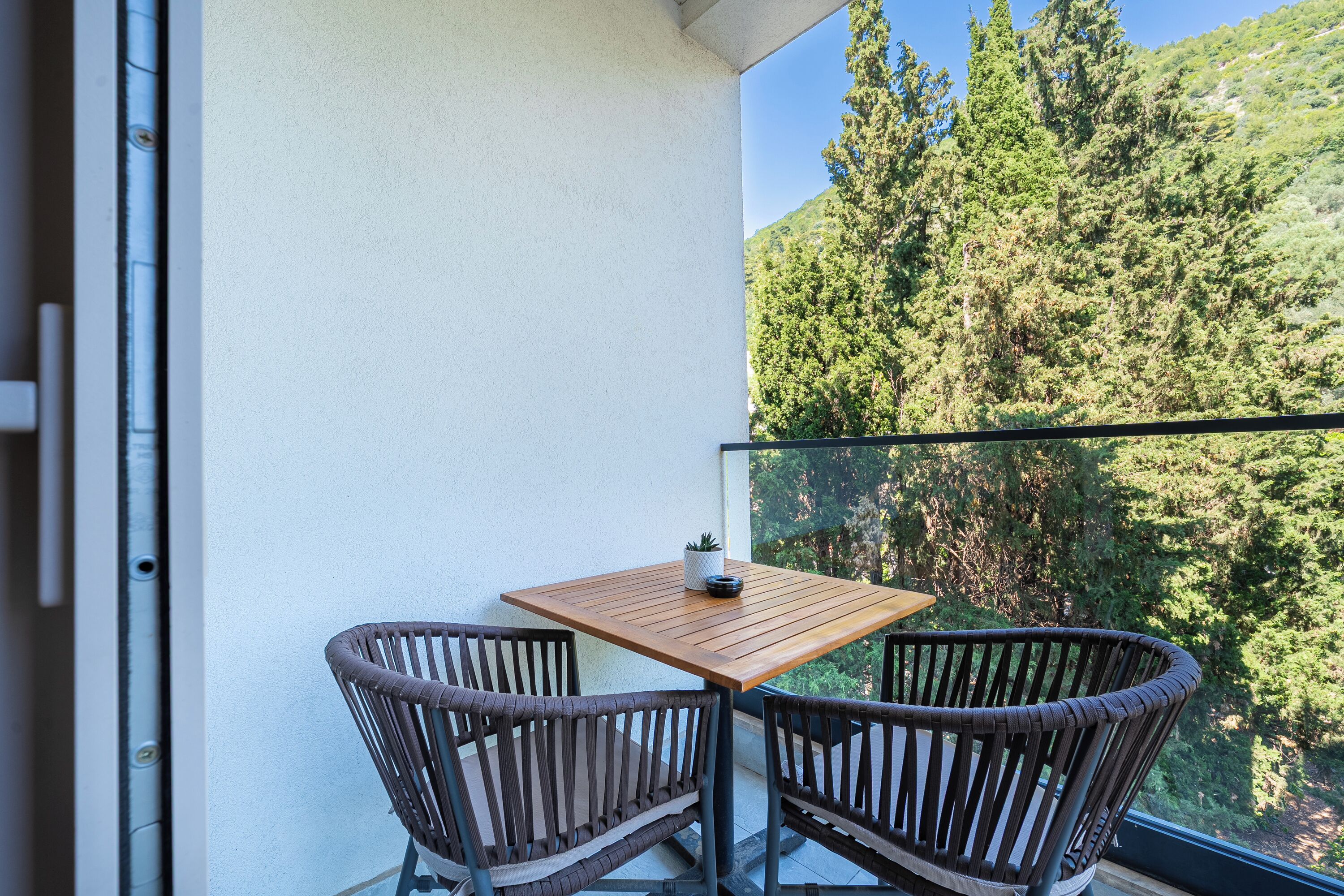 A balcony with a wooden table and wicker chairs, overlooking a forested hillside.