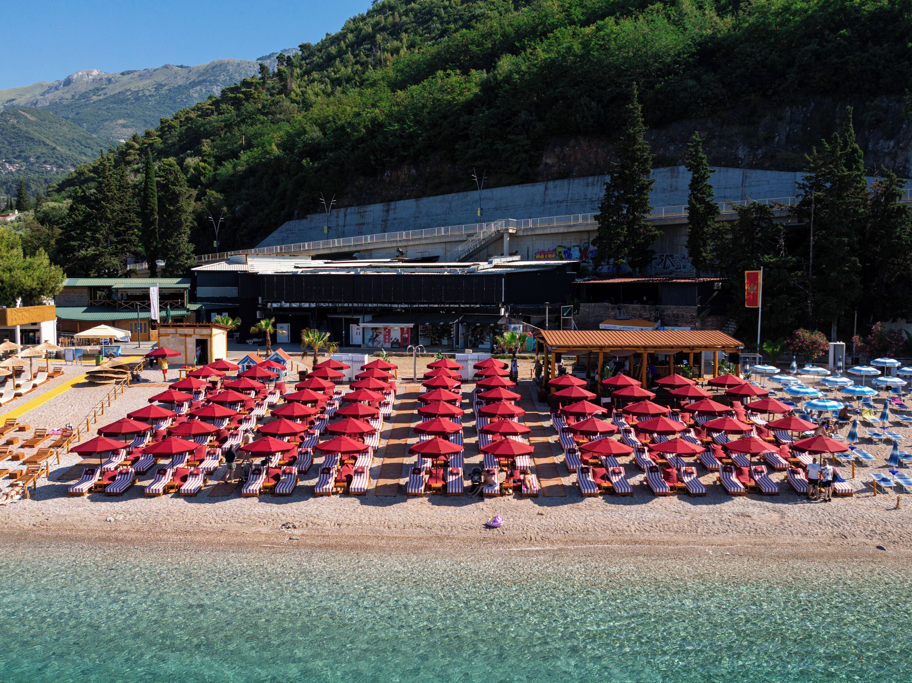Ein Strand mit Reihen von Liegestühlen und Sonnenschirmen, ein Gebäude mit einer Bühne und eine gebirgige Kulisse.