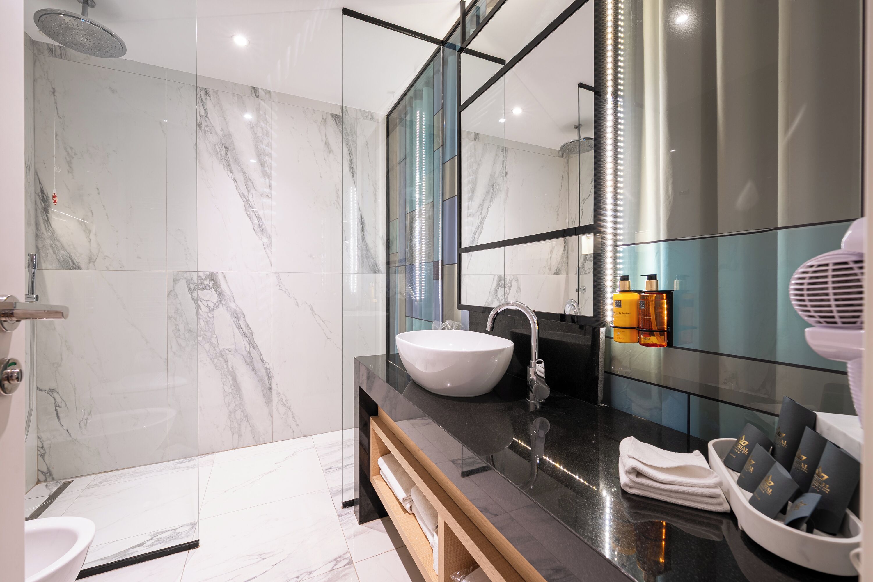 A modern bathroom with a marble wall, a white sink, a glass shower enclosure, and a vanity with toiletries.