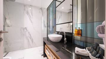 A modern bathroom with a marble wall, a white sink, a glass shower enclosure, and a vanity with toiletries.