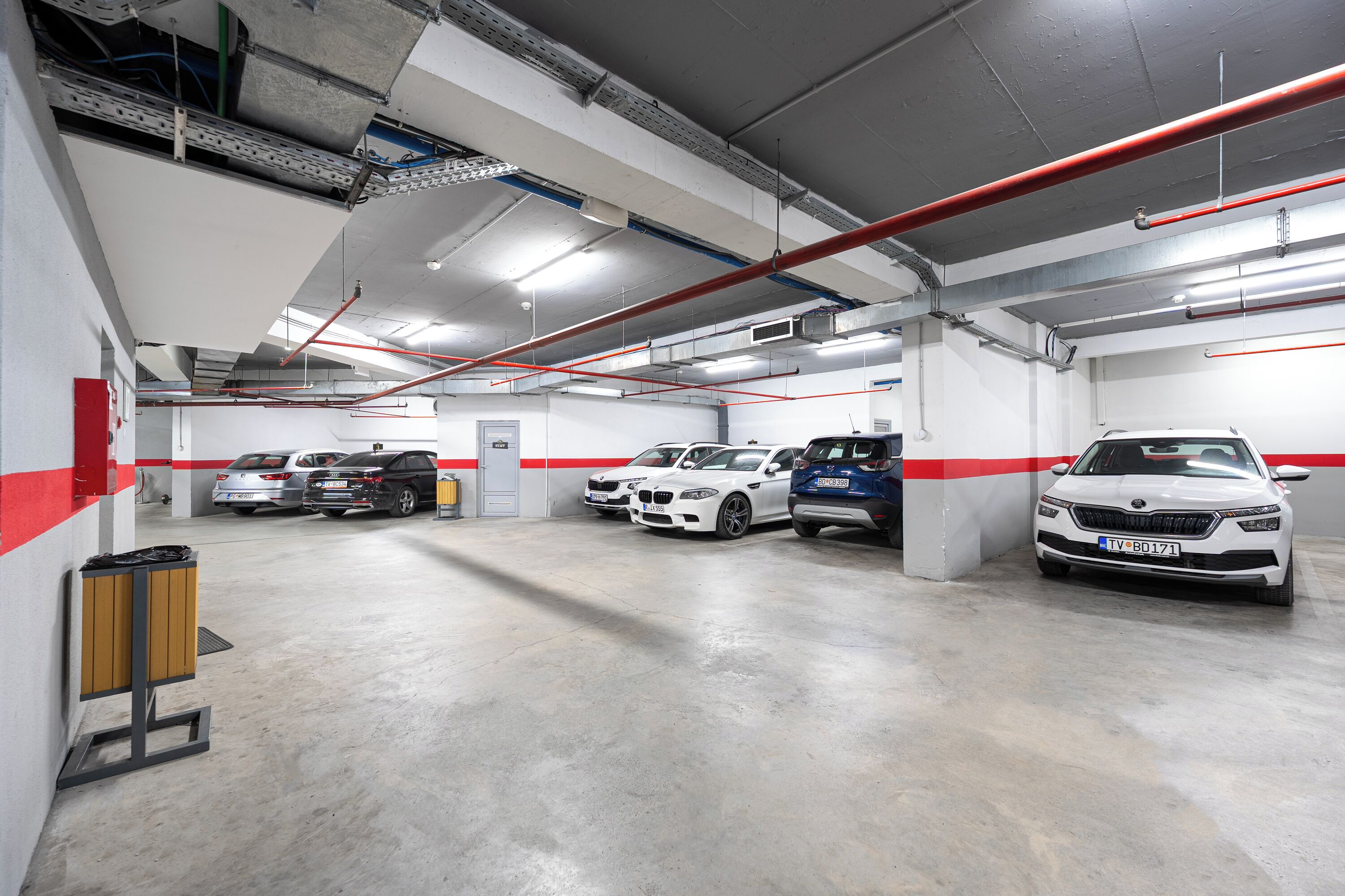 A multi-level parking garage with several cars parked, concrete floors, and visible structural elements like beams and pipes.