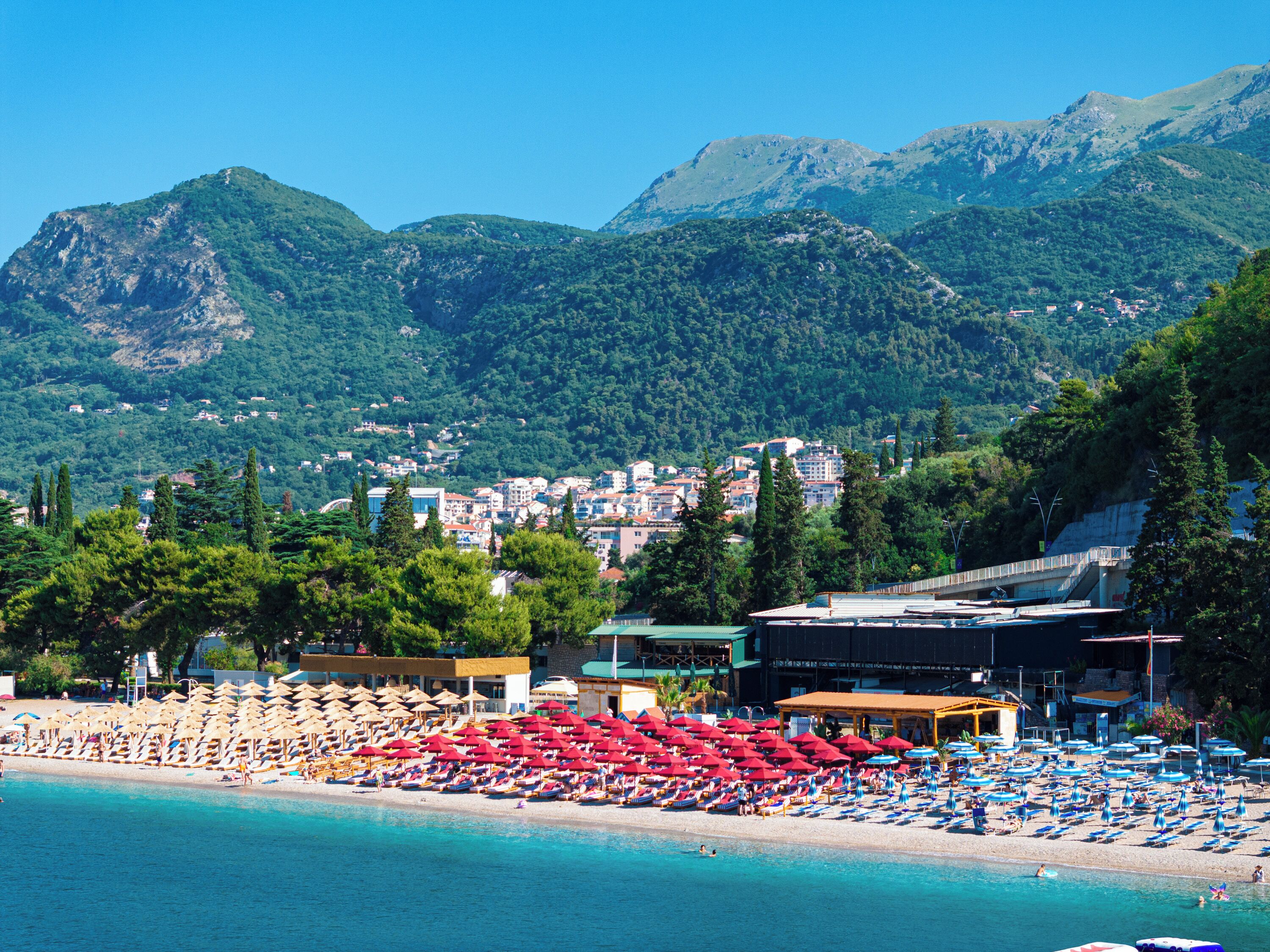 A coastal resort with beach umbrellas, sun loungers, and a view of a town and mountains in the background.
