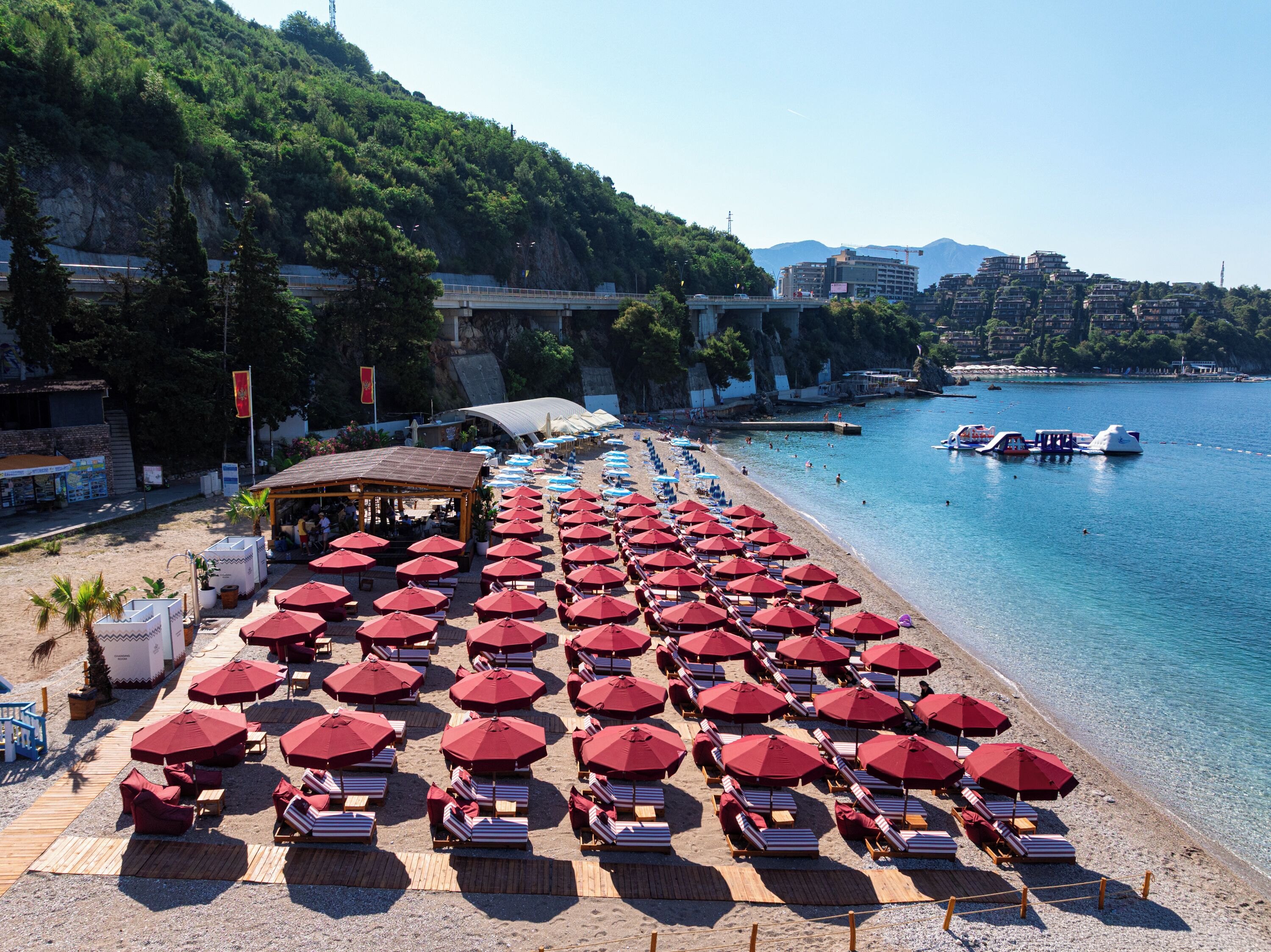 Ein Strand mit Reihen roter Sonnenschirme und Liegestühle, eine Pier, die sich ins Wasser erstreckt, und Gebäude im Hintergrund.
