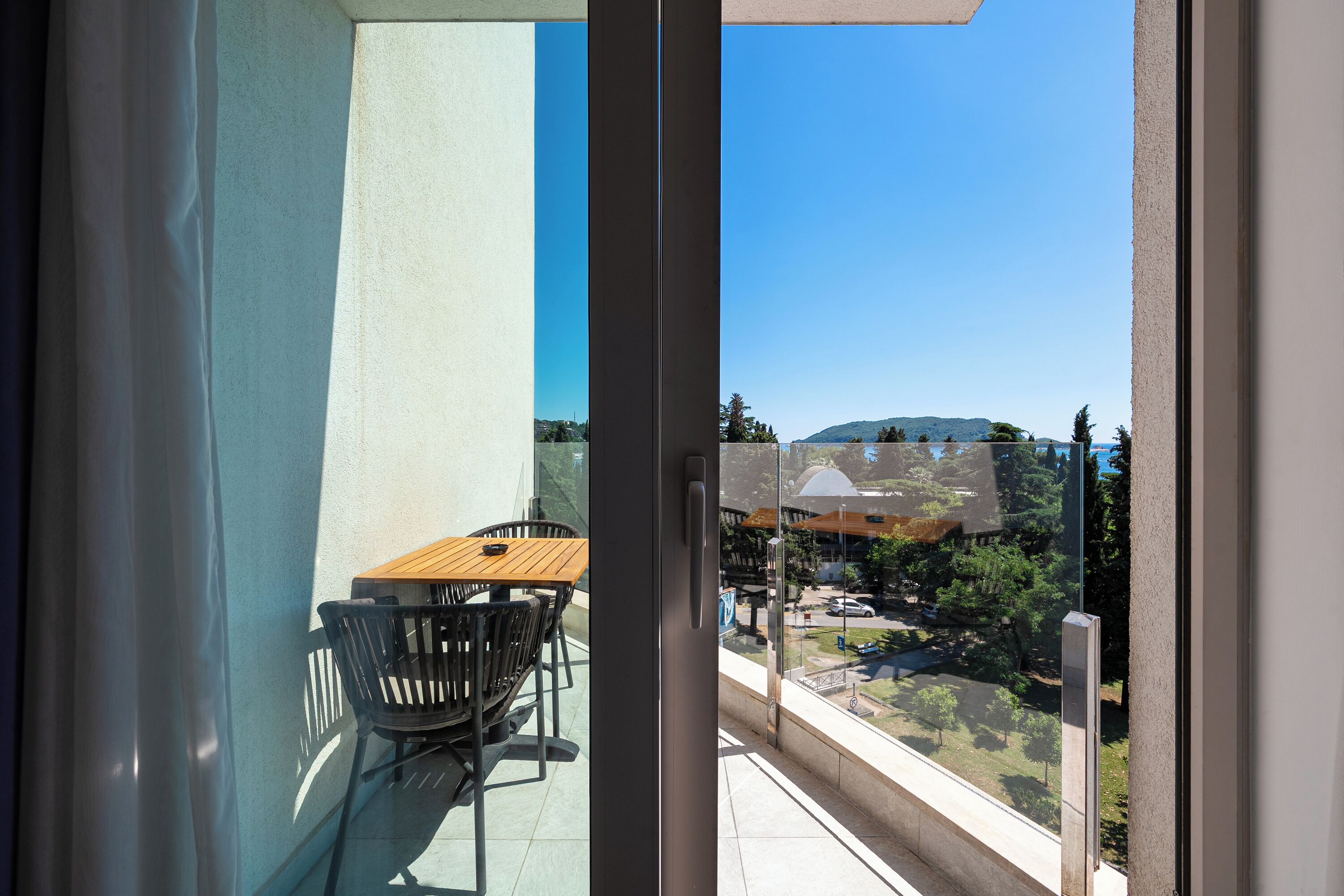 Un balcón con mesa y sillas, un ventanal de vidrio y vistas a un edificio y árboles.