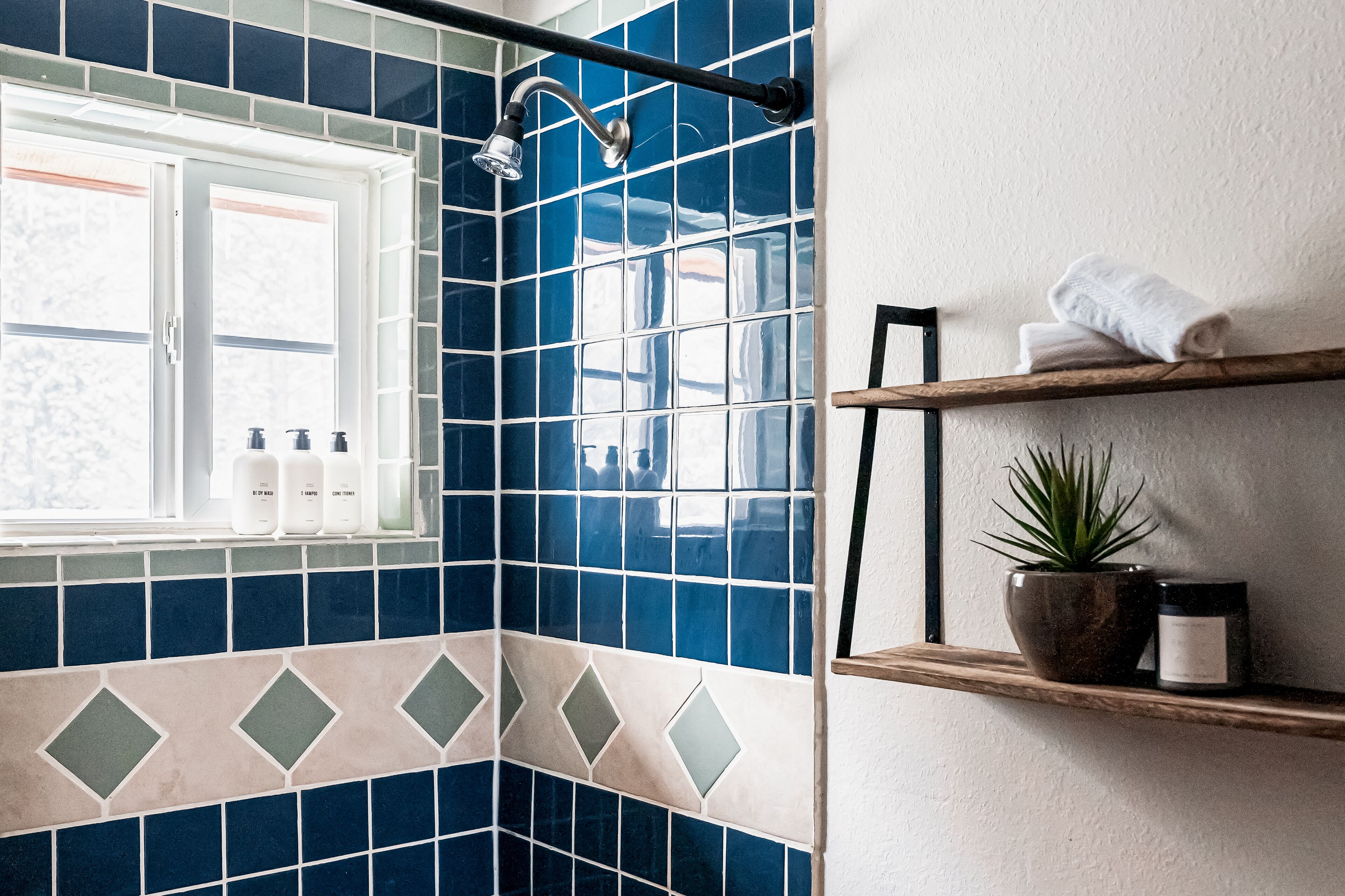 A bathroom with blue and beige tiles, a window, a shower, and shelves with towels and a plant.