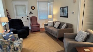 A living room with a patterned armchair, a dark armchair, a grey sofa, and a red chair. There is a rug on the floor, a lamp on a side table, and a clock on the wall. - 1st-floor, 4-bedroom home, 1 block from beach in Ocean City, NJ (Ocean City)