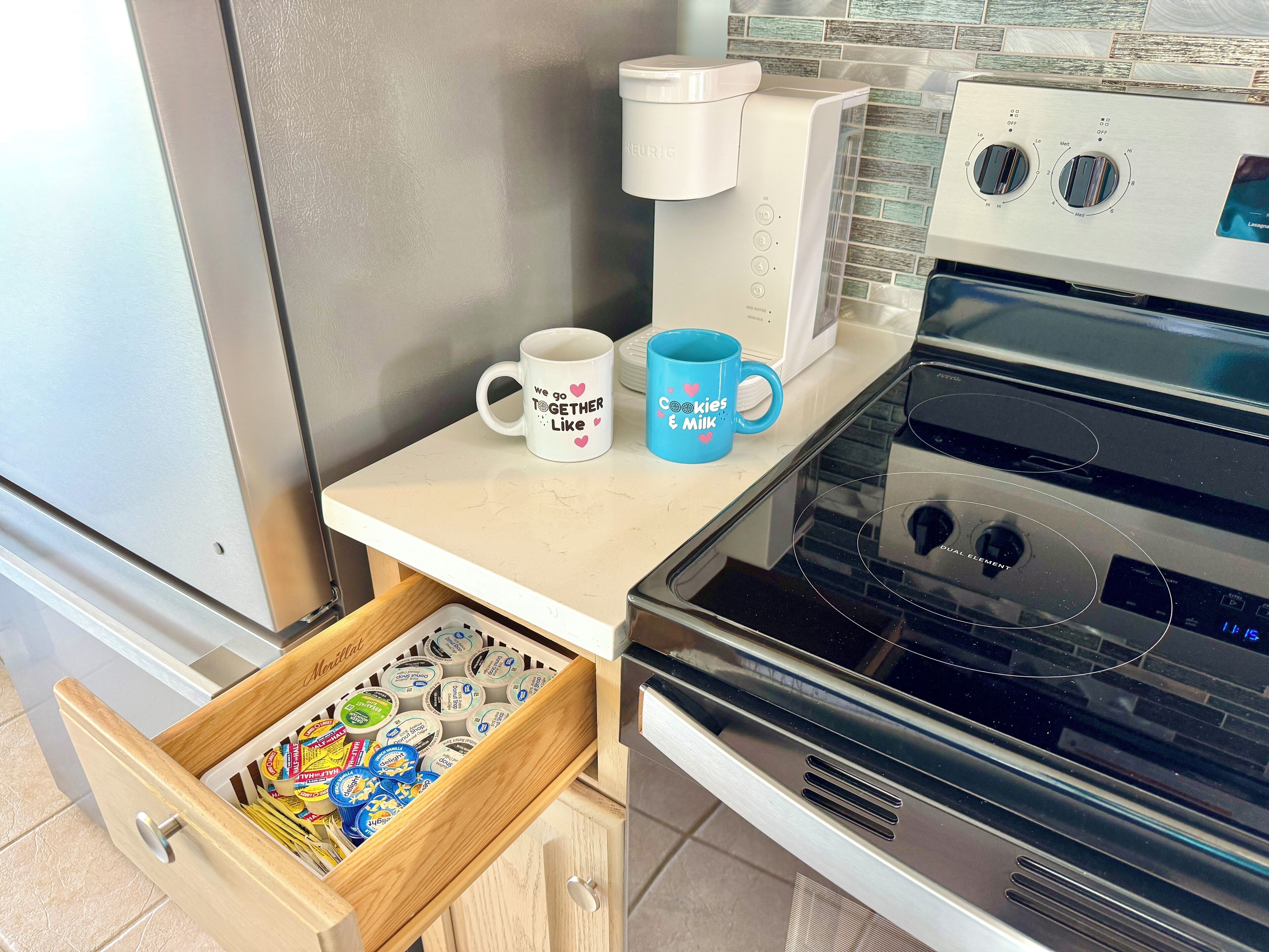 A kitchen with a coffee maker, two mugs, and a drawer with food items.