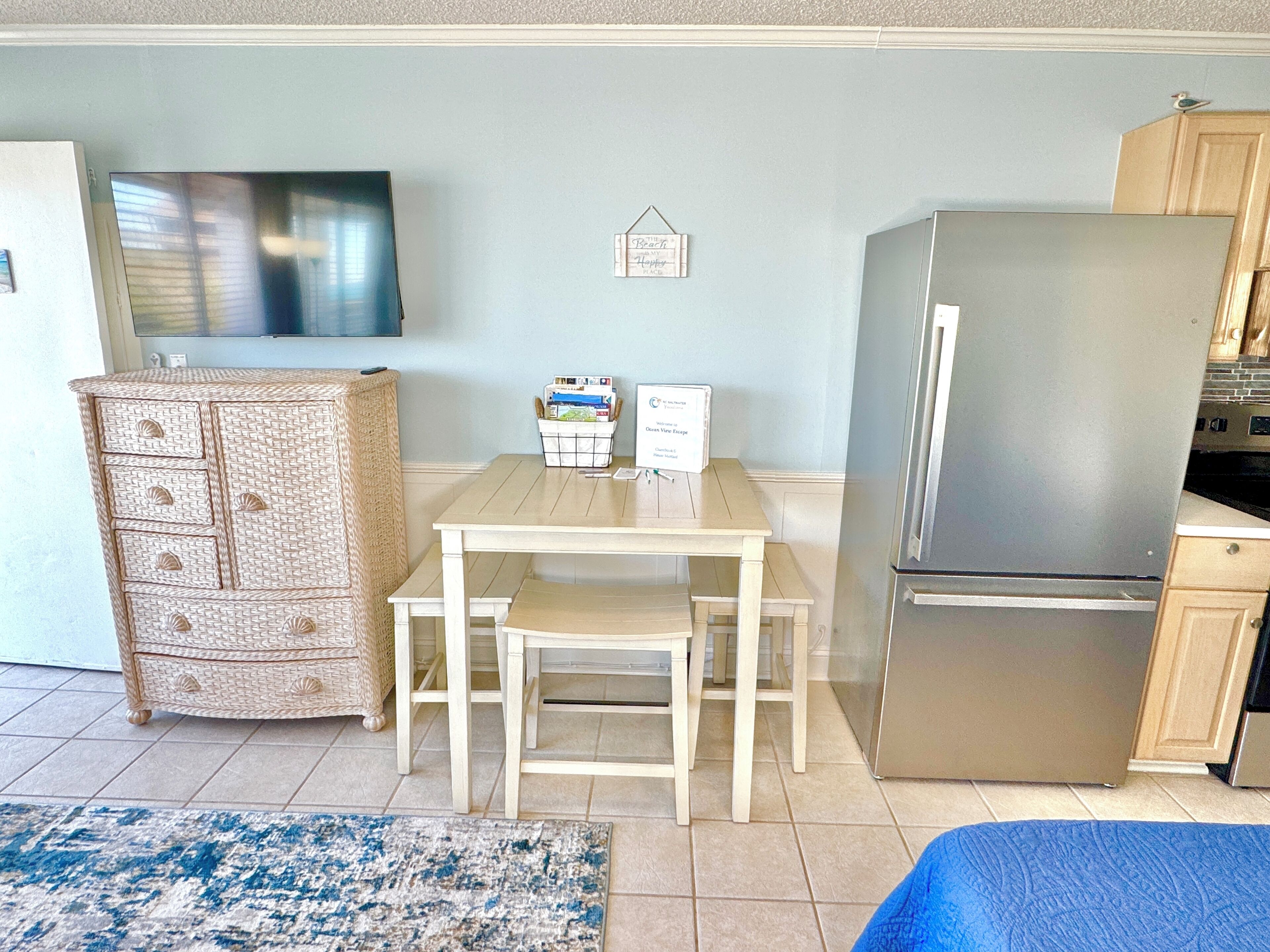 Une cuisine compacte comprenant un réfrigérateur, une table à manger avec des chaises, une commode en osier et un téléviseur fixé au mur.