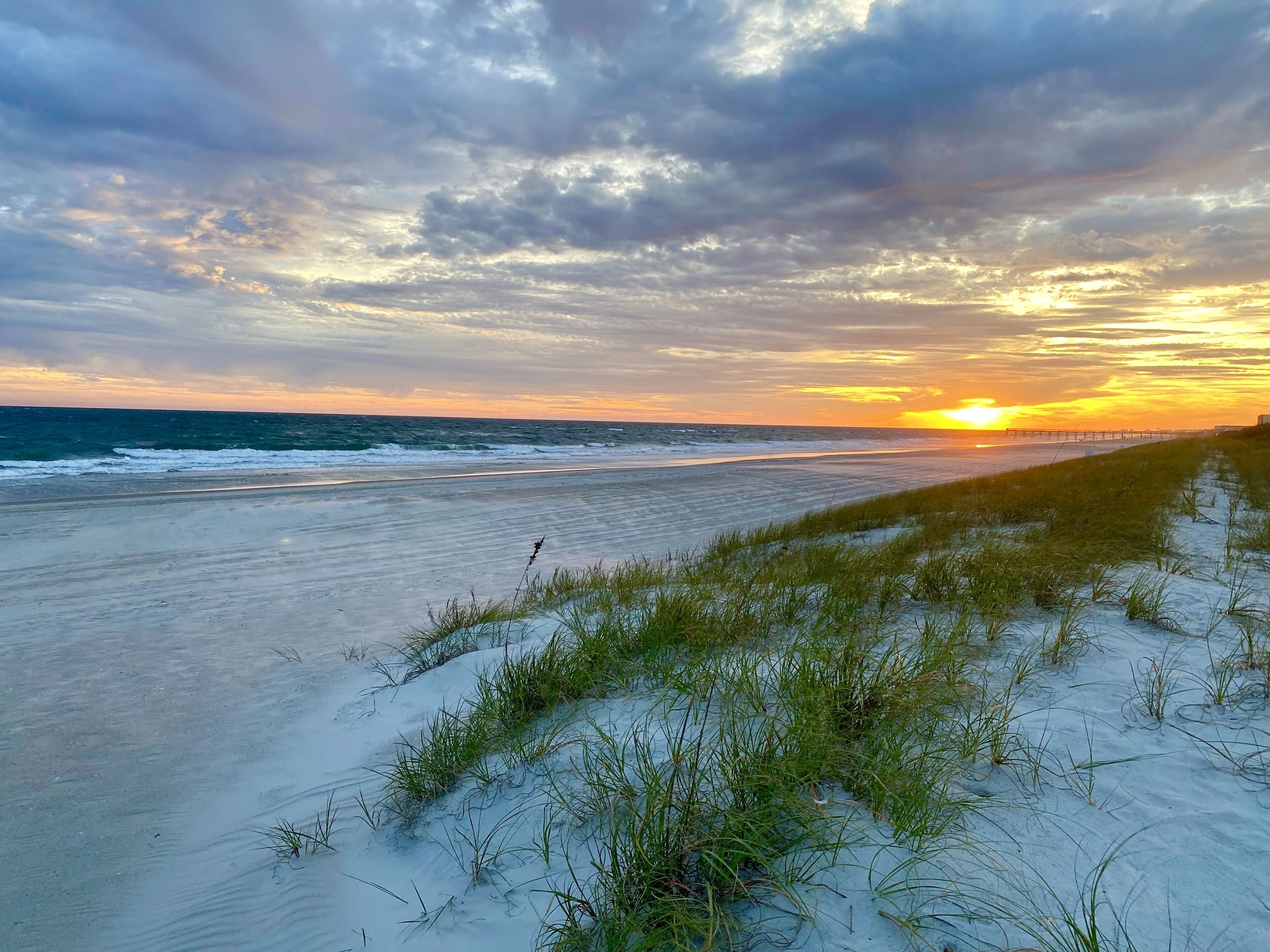 Une plage avec des dunes de sable et de l’herbe au premier plan, une jetée qui s’avance dans l’océan, et un coucher de soleil en arrière-plan.