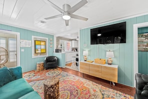 A room with teal walls, a ceiling fan, a black leather chair, a colorful rug, a wooden sideboard, and a kitchen area in the background.