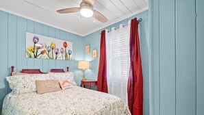A bedroom with a floral bedspread, a ceiling fan, a bedside table with a lamp, and a window with red curtains. - High Tide in Windy Hill (North Myrtle Beach)