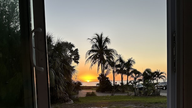 Vista a través de una ventana de un área exterior pavimentada con palmeras y un atardecer de fondo.