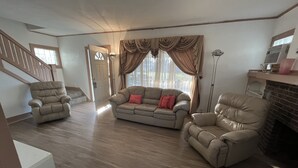 Un salon spacieux avec un canapé beige, deux fauteuils et une cheminée. On aperçoit un escalier au fond de la pièce.